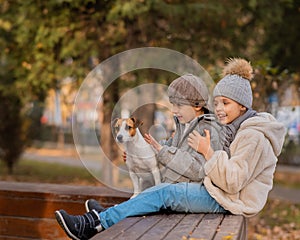 Brother and sister sit in an embrace with a dog on a bench for a walk in the autumn park. Boy, girl and jack russell