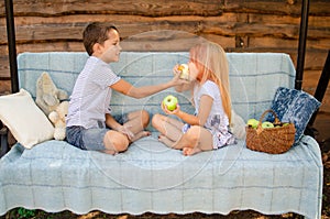 Brother and sister sit opposite each other on a garden swing and eat green apples. Beautiful happy caucasian children in the