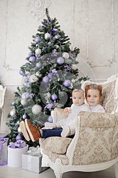 Brother and sister are sitting on a vintage armchair in a white room beautiful decorated for christmas holiday with a Christmas