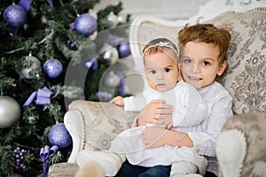 Brother and sister are sitting on a vintage armchair in a white room beautiful decorated for christmas holiday with a Christmas