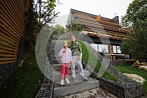 Brother with sister stand on stairs against modern wood stone house