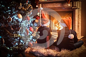 Back view, brother and sister wearing Santa`s hats warming next to a fireplace in a living room decorated for Christmas.