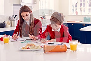 Brother And Sister Wearing School Uniform Doing Homework On Kitchen Counter With Healthy Snacks