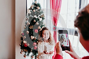 brother taking a picture of his sister with mobile phone at home. Girl standing by the christmas tree. Family time. Christmas