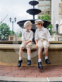 Brothers with bowties and hats sitting on a fountain and looking at each other in a park