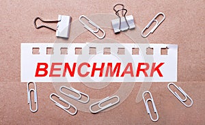 On a brown background, white paper clips and a torn strip of white paper with the text BENCHMARK