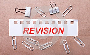 On a brown background, white paper clips and a torn strip of white paper with the text REVISION
