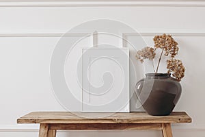 Brown bold vase. Dry hydrangea flowers on old wooden bench. Blank black picture frame mockup. White wall moulding
