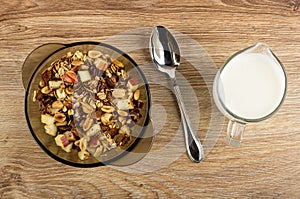 Brown bowl with granola, spoon, pitcher with yogurt on wooden table. Top view