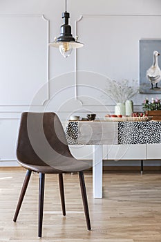 Brown chair under lamp in white dining room interior with table and wall with molding. Real photo