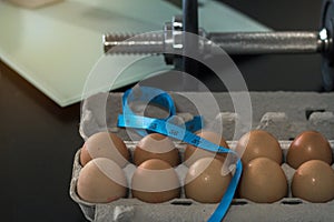 Brown chicken eggs in an open egg carton, dumbbell with weighing scale and measuring tape on black table background.