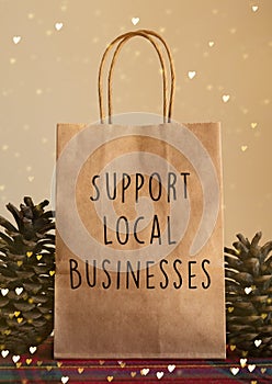 Brown Christmas paper bag with `support local businesses` message in front of a beige background with pine cone props photo