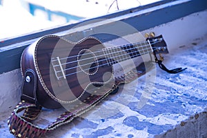 Brown colored ukulele with strap leaning against a brick window frame. Concept instruments, music, strings