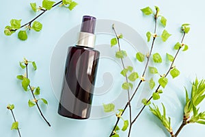 Brown cosmetic spray bottle and birch branches with young small leaves on blue background. Mockup. Skincare beauty and liquid