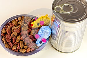 Brown crunchy organic kitty kibble pieces in a bowl, toy mice and wet cat food in metal can for pet feed on light background.