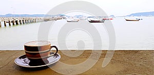 Brown cup of coffee on concrete table with seascape, ocean, bridge and boat background.