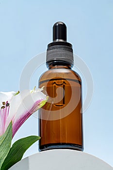 A brown dropper bottle of face oil displayed on a pedestal against a blue background with pink alstromeria flower. cosmetic