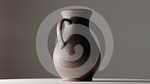 Brown earthenware jug with a handle spinning in a dark studio. An old jar from clay rotating on a white table isolated