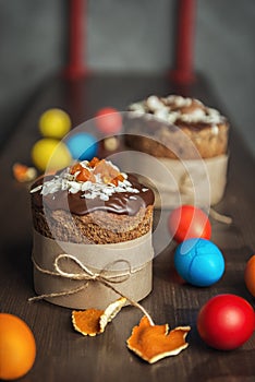 Brown Easter cakes decorated with chocolate, almond flakes and dried apricots among colored Easter eggs on wooden table