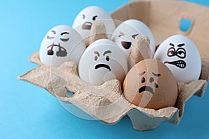 Brown egg with upset face among aggressively disposed white ones in carton box on turquoise background, closeup. Bullying concept