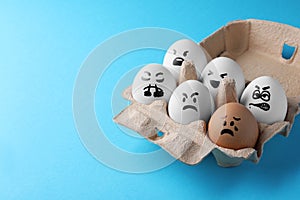 Brown egg with upset face among aggressively disposed white ones in carton box on turquoise background, space for text. Bullying