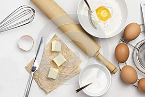 Brown eggs on metal stand. Butter and knife on paper. Egg yolk with flour and spoon in bowl. Milk in bowl and rolling pin on table