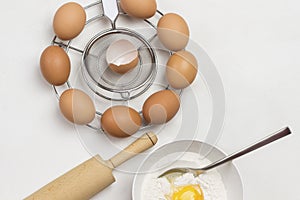 Brown eggs on metal stand. Egg yolk with flour and spoon in bowl. Milk in bowl and rolling pin on table