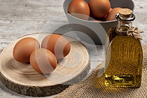 Brown eggs on a piece of wood, next to a bottle of virgin olive oil and in the background more eggs in a gray bowl, all on a