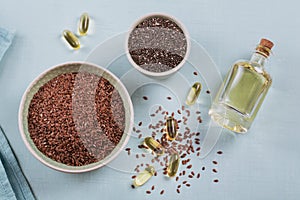 Brown flax seed or linseed and chia in small bowl and gelatin capsules with omega oil on light blue background