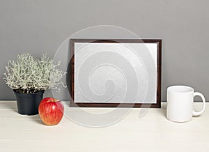 Brown frame mockup with plant pot, mug and apple on wooden shelf