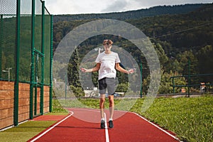 Brown-haired boy with an athletic figure wearing a white T-shirt and black shorts is jumping rope on an athletic oval. Training to