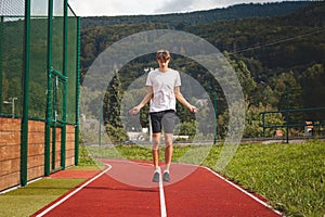 Brown-haired boy with an athletic figure wearing a white T-shirt and black shorts is jumping rope on an athletic oval. Training to