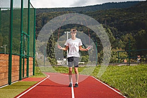 Brown-haired boy with an athletic figure wearing a white T-shirt and black shorts is jumping rope on an athletic oval. Training to