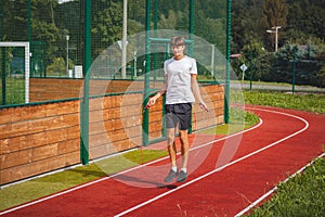 Brown-haired boy with an athletic figure wearing a white T-shirt and black shorts is jumping rope on an athletic oval. Training to