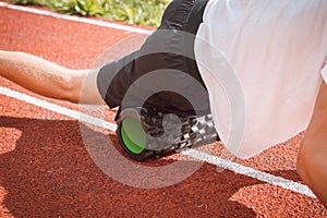 Brown-haired boy with an athletic physique on an athletic oval massages his muscles with a foam roller for better recovery. Post-