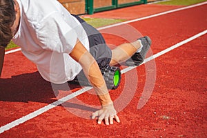 Brown-haired boy with an athletic physique on an athletic oval massages his muscles with a foam roller for better recovery. Post-