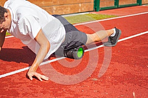 Brown-haired boy with an athletic physique on an athletic oval massages his muscles with a foam roller for better recovery. Post-