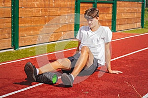 Brown-haired boy with an athletic physique on an athletic oval massages his muscles with a foam roller for better recovery. Post-