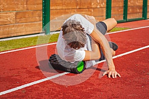Brown-haired boy with an athletic physique on an athletic oval massages his muscles with a foam roller for better recovery. Post-