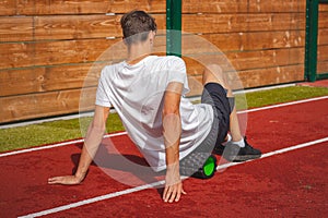 Brown-haired boy with an athletic physique on an athletic oval massages his muscles with a foam roller for better recovery. Post-