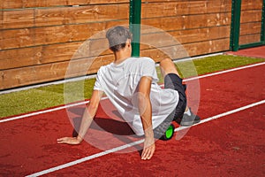 Brown-haired boy with an athletic physique on an athletic oval massages his muscles with a foam roller for better recovery. Post-