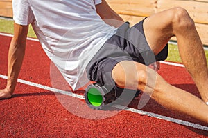 Brown-haired boy with an athletic physique on an athletic oval massages his muscles with a foam roller for better recovery. Post-