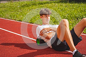 Brown-haired boy with an athletic physique on an athletic oval massages his muscles with a foam roller for better recovery. Post-