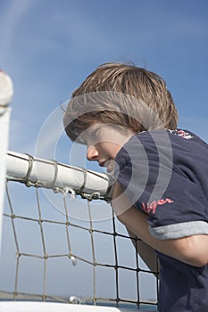 Brown-haired boy in t-shirt, hanging over the railing of a ferry and looking around, in the background a little rainbow