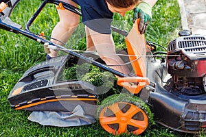 brown haired female gardener raking fresh grass out lawn mower or grass cutter in garden. Agriculture