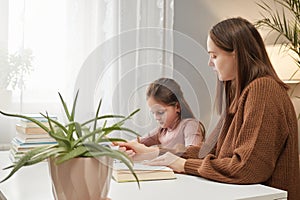 Brown haired mother and daughter learning to write woman teaches child helps with homework while sitting at table in living room