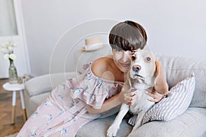 Brown-haired smiling girl in pink dress playing with her cute beagle dog at home. Indoor portrait of lovely young lady