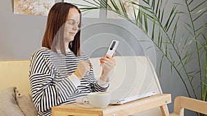 Brown haired woman wearing casual shirt sitting on sofa with closed laptop and coffee, using her smartphone while having break