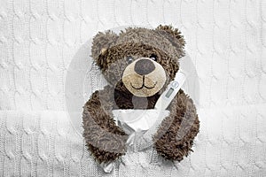 Brown ill teddy bear with thermometer in white bedroom