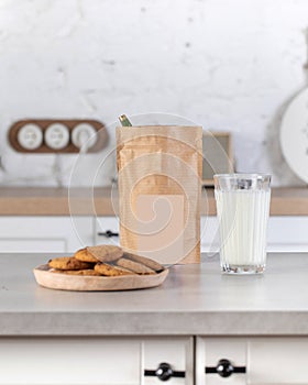 A brown kraft paper bag with a label layout on the kitchen table with a plate of cookies and a glass of milk.